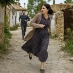 A woman runs away down a village street while looking back.