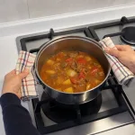 Hands using a simple hand-woven cloth potholder to lift a pot from a stove.