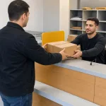 Adult handing a cardboard package to a clerk at an unbranded cargo office counter.